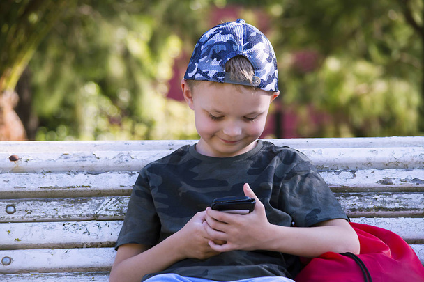 cute boy looking mobile phone on bench in summer - Photo, Image