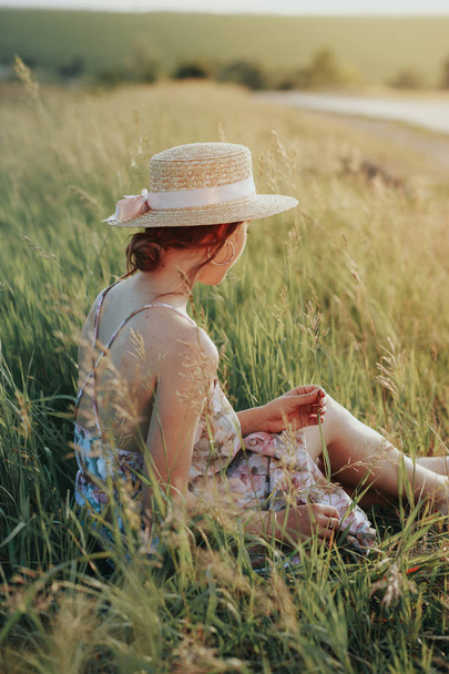 Young girl in a straw hat sits on the grass, holds a flower in the hands, a basket on the side - Valokuva, kuva