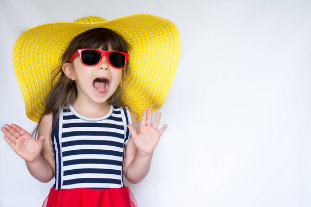 Χαρούμενη έκπληξη κοριτσάκι σε πολύχρωμα ρούχα και τα γυαλιά ηλίου που στέκεται πάνω σε λευκό φόντο στο. Παιδική μόδα. Καλοκαιρινές διακοπές. - Φωτογραφία, εικόνα
