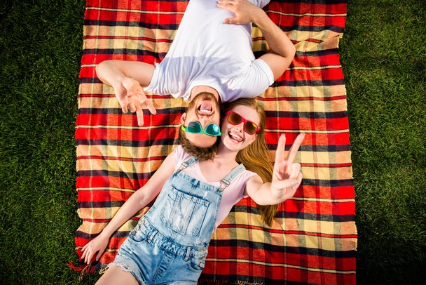 Retrato de vista superior de alegre pareja funky acostada sobre hierba verde cabeza a cabeza mostrando v-signo con dos dedos disfrutando de vacaciones de fin de semana vacaciones teniendo placer
 - Foto, Imagen