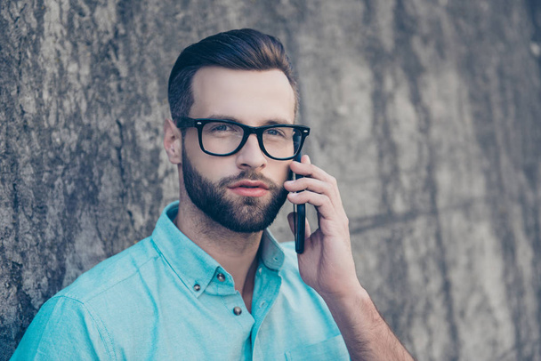 Head shot portrait of cool harsh man with modern hairdo having important business call looking away using smart phone isolated on grey stone wall. Technology gadget concept - Photo, Image