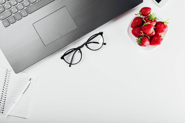 Vista superior del espacio de trabajo freelancer con laptop, notebook, smartphone, gafas y snack. Tener fresas mientras se trabaja. Poner notas
 - Foto, Imagen