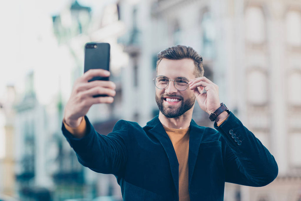 Portrait of cheerful joyful man in eyewear jacket holding eyelet of glasses on face shooting selfie on front camera of smart phone isolated on blurred background. Vacation weekend holiday concept - Fotó, kép