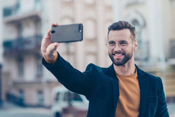 Portrait of cheerful glad gentleman in eyewear jacket shooting selfie on front camera of smart phone isolated on blurred background. Electronic wireless device apps concept - Φωτογραφία, εικόνα