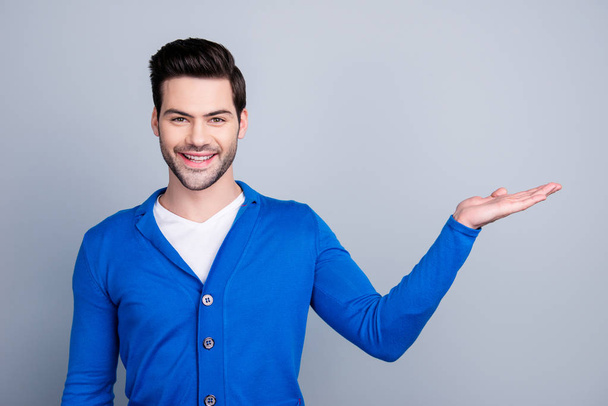 Portrait of positive guy showing copy space on his palm, looking at camera, dressed in blue cardigan, standing over grey background - Fotoğraf, Görsel
