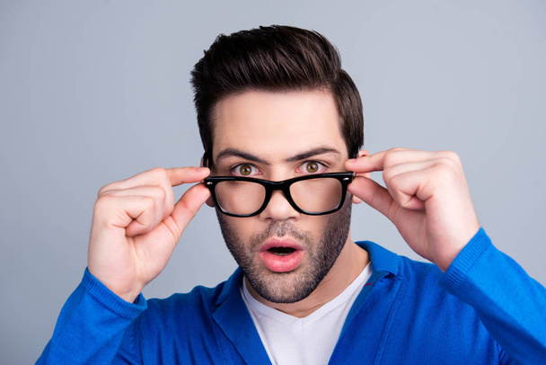 Portrait of amazed man looking out glasses, holding eyelets with two hands, having open mouth, isolated on grey background, dressed in blue pullover - Фото, изображение