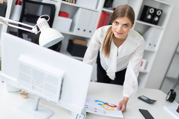 Una joven está de pie doblada cerca de una mesa en la oficina. La chica trabaja con un ordenador, bloc de notas y documentos
. - Foto, Imagen