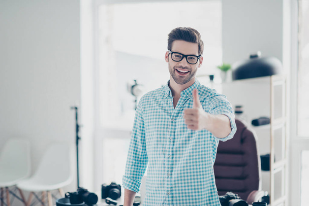 Portrait of positive cheerful guy in shirt with stubble and modern hairstyle standing in photo studio, laughing,  gesturing thumb up to the camera - Photo, Image