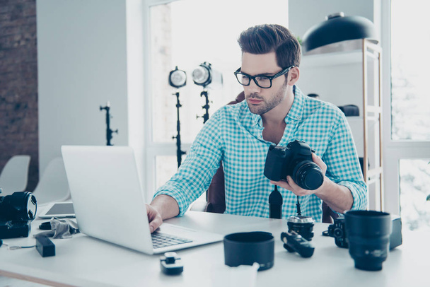 Portrait of smart thoughtful man holding digital camera in hand, searching information on computer, sitting at desk, having electronic devices on the table - 写真・画像