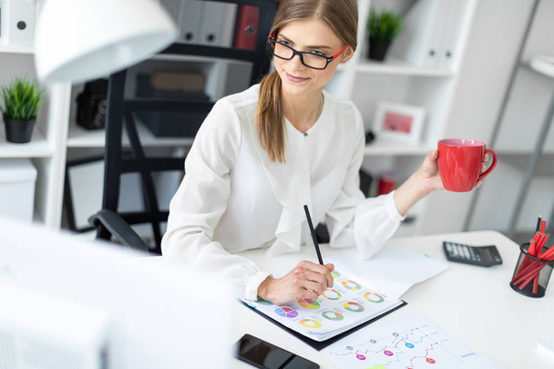 Μια νεαρή κοπέλα κάθεται σε ένα γραφείο υπολογιστή στο γραφείο, κρατώντας ένα μολύβι και ένα φλιτζάνι κόκκινο. Πριν το κορίτσι υπάρχουν διαγράμματα. - Φωτογραφία, εικόνα