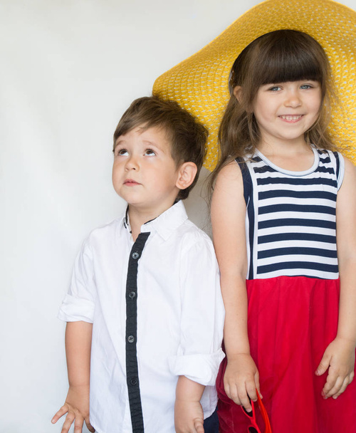 Leuke mode kinderen staan samen. Klein meisje in stro hoed en gestreepte rode jurk en kleine jongen in wit overhemd in de studio op een witte achtergrond. Vakantie, vriendschap, mode voor kinderen - Foto, afbeelding