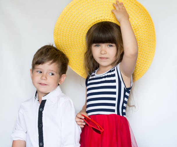 Niedliche Mode-Kids stehen zusammen. kleines Mädchen mit Strohhut und gestreiftem rotem Kleid und kleiner Junge im weißen Hemd im Studio auf weißem Hintergrund. Urlaub, Freundschaft, Mode für Kinder - Foto, Bild