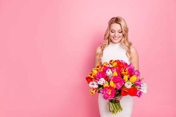 Portrait with copy space empty place of charming pretty woman in white dress having big bouquet of tulips in hands looking at flowers isolated on pink background - Foto, afbeelding