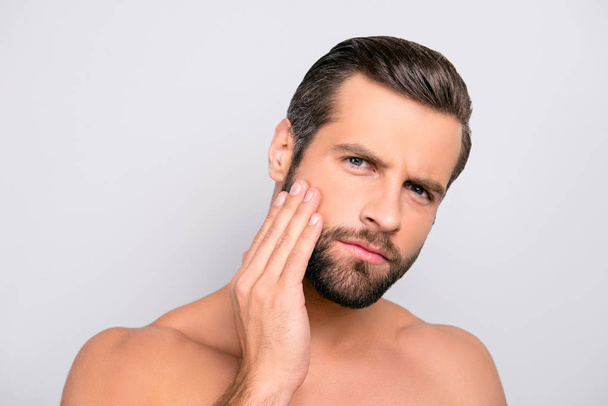 Attractive, unhappy, displeased, harsh, virile macho having problem with face, holding hand on cheek, he needs lotion, cream, mask to make soft, smooth, clear, clean skin, isolated on gray background - Фото, изображение