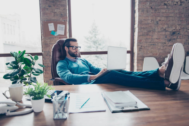 Stunning man in shirt, pullover, jeans sitting on leather chair putting legs on desktop in work station, having computer on his knees with serious expression working on new project - 写真・画像