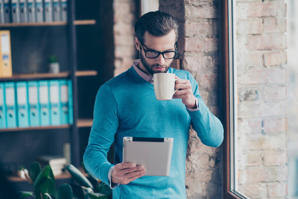 Portrait of concentrated confident clever intelligent busy banker dressed in formalwear blue pullover checkered shirt enjoying tea reading new using digital tablet standing near window at workstation - Foto, imagen