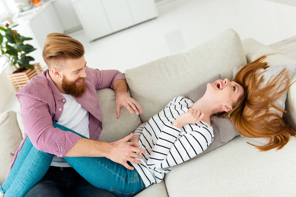 Entertainment fun-time wife husband living room spouse two partners concept. Cute funky lovely cheerful excited adorable pretty lady lying on divan covering closing body with hands afraid of tickle - Fotoğraf, Görsel