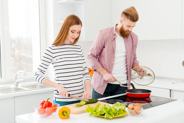 Breakfast supper dinner kitchen ware concept. Portrait of busy concentrated partners cooking meal together, beautiful woman cutting cucumber handsome man frying food in modern white kitchen - Photo, image