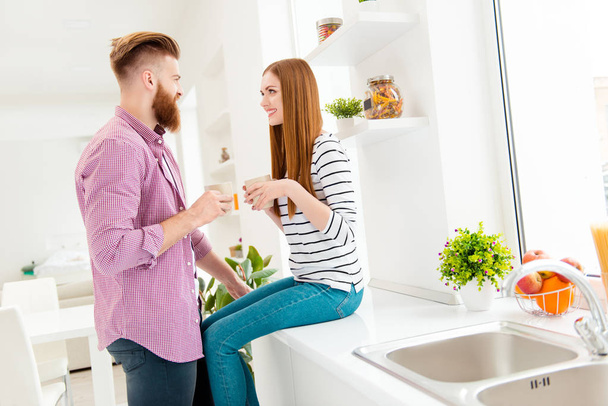 Weekend holiday vacation pleasure conversation concept. Side view portrait of lovely romantic couple holding cups with hot beverage in hands enjoying morning together in modern white kitchen - Photo, image