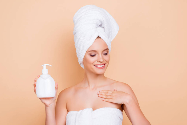 Portrait of cheerful positive model with turban on head after shower applying body lotion from white bottle with dispenser enjoying aroma isolated on beige background - Photo, image
