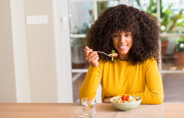 Afrikalı-Amerikalı kadın ayakta ve diş gösterilen kendine güvenen bir gülümsemeyle gülümseyerek mutlu bir yüz ile makarna salatası evde yemek - Fotoğraf, Görsel