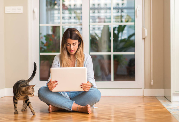 Nuori kaunis nainen hymyilee istuu lattialla tietokoneen ja kissan kanssa
. - Valokuva, kuva
