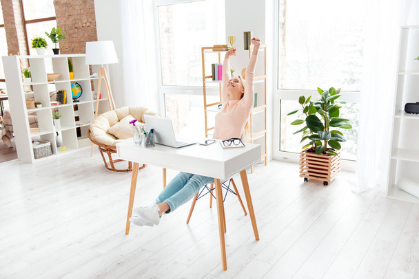 Portrait of cheerful positive girl sitting in white room at desktop making stretching with raised arms while preparing for exams enjoying timeout dreaming about weekend holiday - Foto, imagen