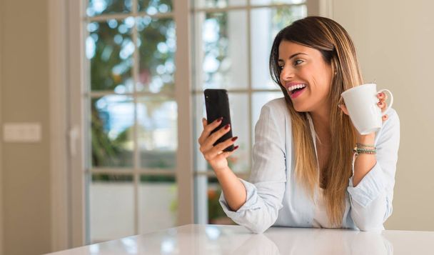 Όμορφη νεαρή γυναίκα κοιτάζοντας το τηλέφωνο και χαμογελώντας ενώ πίνουν καφέ στο σπίτι. - Φωτογραφία, εικόνα