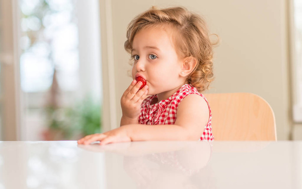 Hermosa niña rubia con ojos azules comiendo fresa en casa
. - Foto, imagen