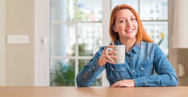 rothaarige Frau hält eine Tasse Kaffee mit einem glücklichen Gesicht im Stehen und lächelt mit einem selbstbewussten Lächeln, das Zähne zeigt - Foto, Bild
