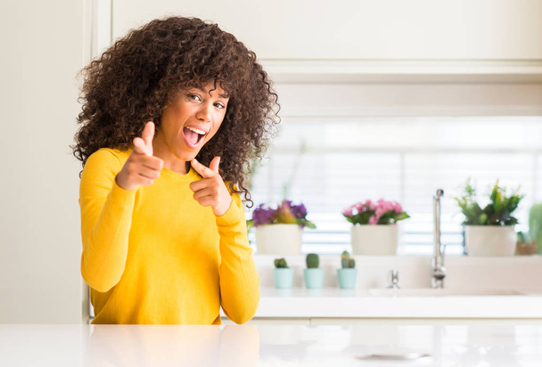 Afrikkalainen amerikkalainen nainen yllään keltainen pusero keittiössä osoittaa sormella kameraan onnellinen ja hauska kasvot. Hyvä energia ja vibes
. - Valokuva, kuva