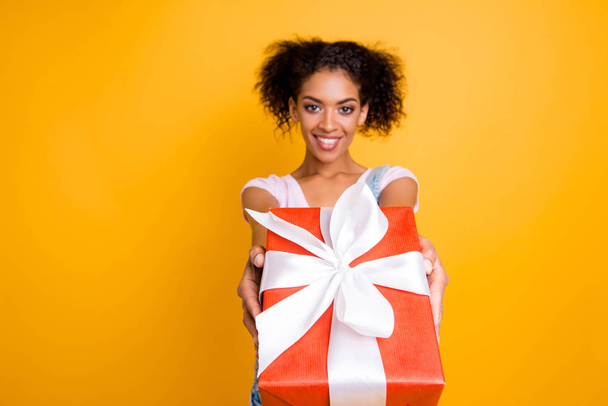 Portrait of friendly positive girl giving front big gift box in red package with white bow isolated on yellow background - 写真・画像