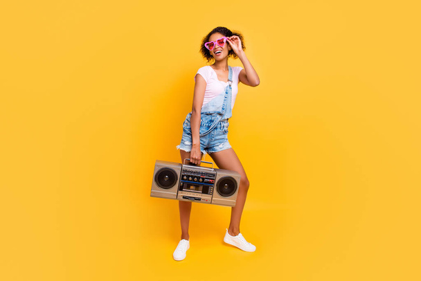 Full size portrait of cheerful positive girl in eyewear jeans overall holding boom box in hand going to make party isolated on yellow background. Music lover fan concept - Fotografie, Obrázek