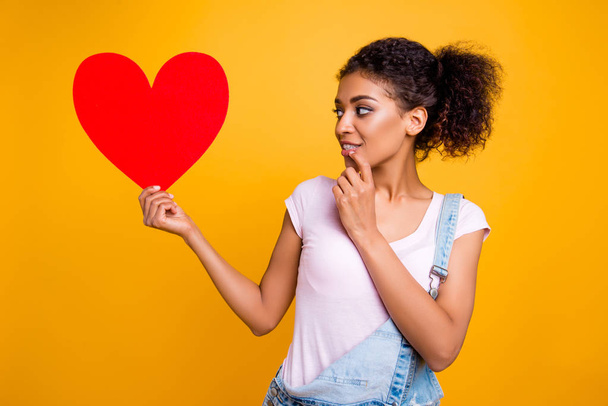 Portrait of thoughtful dreamy girl having big carton paper heart in hand looking at love symbol holding hand on chin isolated on yellow background - Zdjęcie, obraz
