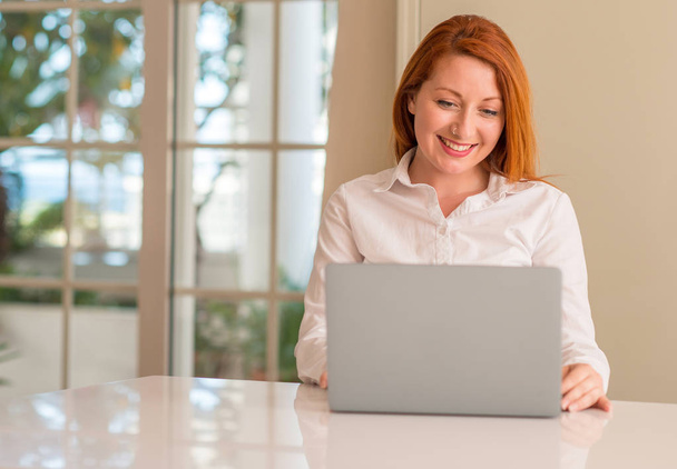 Roodharige vrouw met behulp van computer laptop thuis met een blij gezicht permanent en lachend met een vertrouwen glimlach weergeven tanden - Foto, afbeelding
