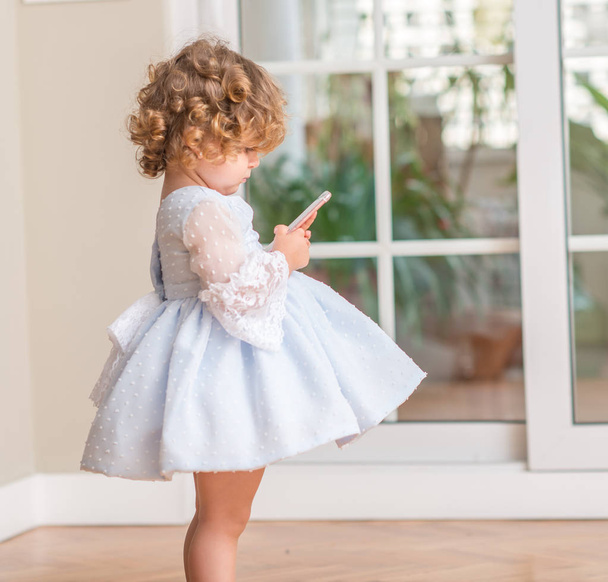 Beautiful blonde child in a dress standing playing with smartphone at home. - Zdjęcie, obraz