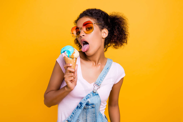 Portrait of childish funky girl in eyewear eyeglasses having ice cream in waffle cone gesturing tongue out isolated on yellow background. Mood style lifestyle concept - Foto, Imagem