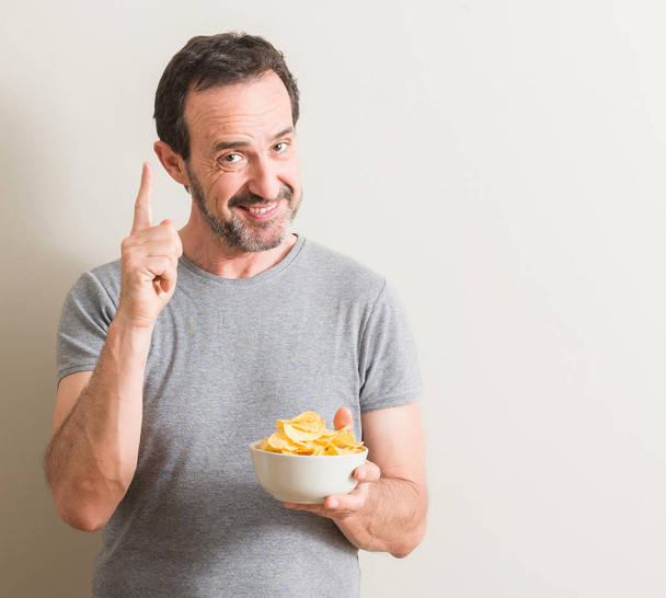 Senior man eating potato chips surprised with an idea or question pointing finger with happy face, number one - Photo, Image