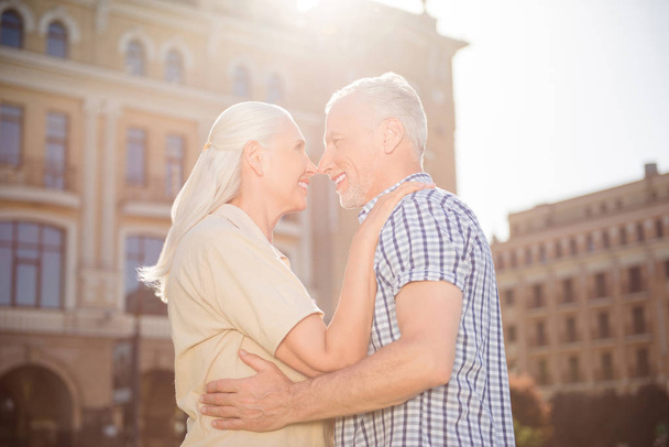 Side view portrait of beautiful attractive spouses hugging nose to nose standing over blurred buildings outdoor in sunshine enjoying time together. Idyllic concept - Foto, Bild