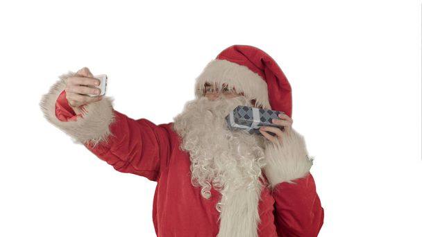 Santa Claus holding a big present  doing a selfie on smartphone on white background - Zdjęcie, obraz