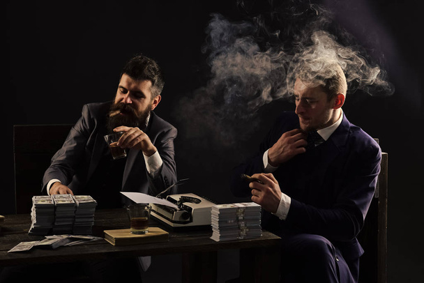 gangster meeting. Illegal business concept. Businessmen discussing illegal deal while drinking and smoking, dark background. Company engaged in illegal business. Men sitting at table with piles of - Foto, Bild