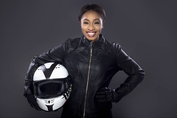 Black female motorcycle biker or race car driver or stuntwoman wearing leather racing suit and holding a protective helmet.  She is standing confidently in a studio - Foto, immagini