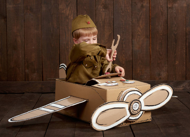 хлопчик одягнений солдатом у ретро військову форму ремонтує літак з картонної коробки, фон з темного дерева, ретро стиль
 - Фото, зображення