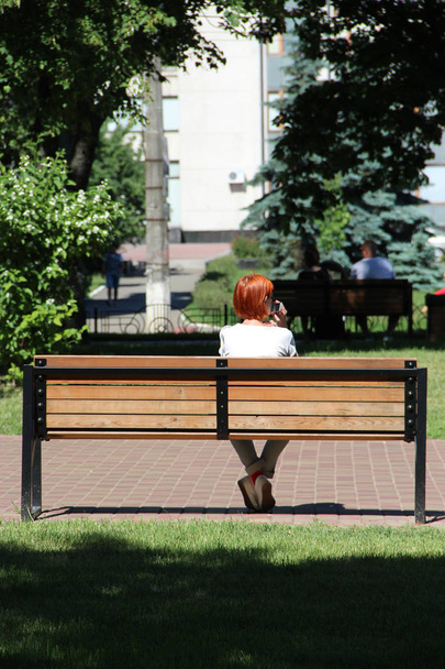 Redhead girl sitting on bench in city park and talking on phone. Young woman with red hairs sitting on bench in park and talking on smartphone. Girl relaxing. Woman has a rest - Photo, Image