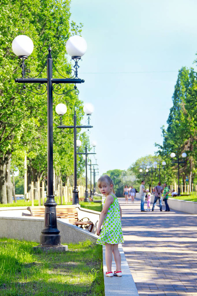 Маленькая девочка, прогуливающаяся вдоль парапета в городском парке и оглядывающаяся в изумлении. Ребенок потерялся во время ходьбы. Детские выражения. Детские эмоции
 - Фото, изображение