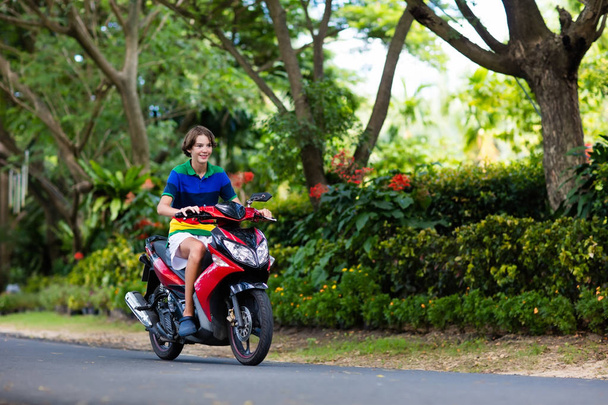 Подросток на скутере. Мальчик-подросток веселится на мотоцикле по дороге в школу. Студент на мотоцикле в тропическом городе. Транспорт и путешествия для молодых взрослых. Путешествуя по городу на велосипеде
. - Фото, изображение