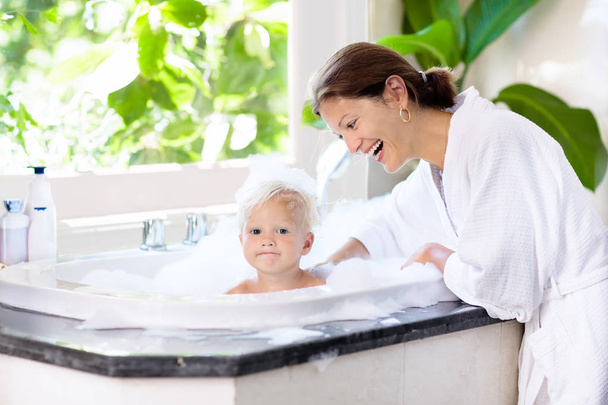 Little child taking bubble bath in beautiful bathroom with big garden view window. Mother washing baby. Kids hygiene. Shampoo, hair treatment and soap foam for children. Mom bathing kid in large tub.  - Foto, immagini
