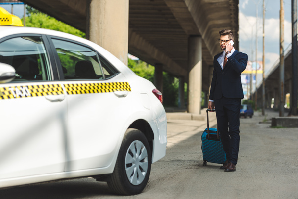 handsome young man in suit and eyeglasses talking by smartphone while standing with suitcase near taxi - Foto, immagini