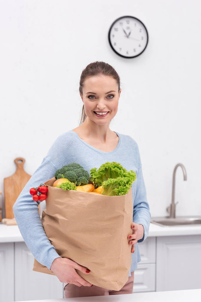 happy adult woman carrying paper bag with vegetables and fruits at kitchen - Foto, immagini