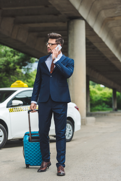 tyylikäs nuori mies puhuu älypuhelimella ja katselee pois seistessään matkalaukku lähellä taksi
 - Valokuva, kuva